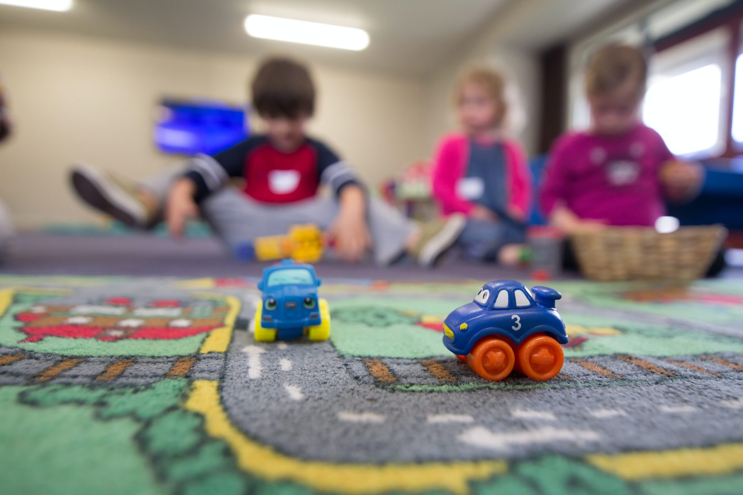 3 preschoolers play on the floor with toy vehicles. The colorful rug shows a road with 2 toy trucks on it.